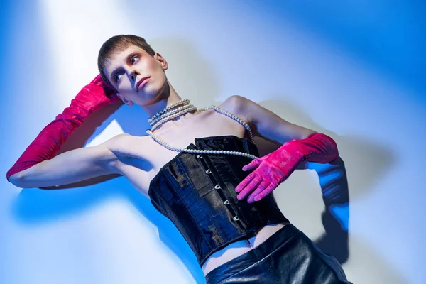 Vista superior, modelo queer en corsé y guantes de color rosa mirando hacia otro lado y acostado sobre fondo azul, genderfluid - foto de stock