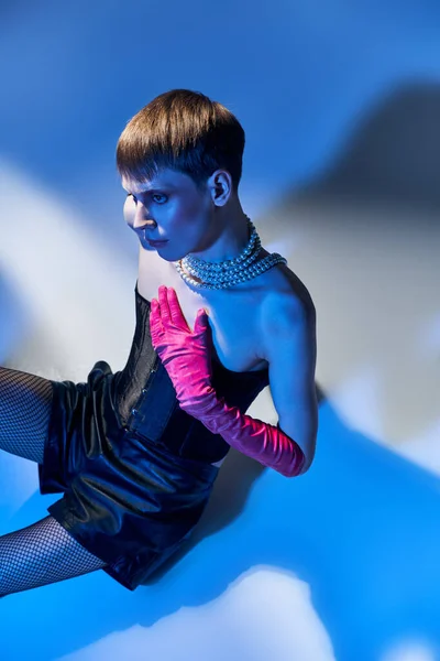 Vista superior, modelo de género fluido en corsé y guantes de color rosa sentado sobre fondo azul, lgbtq, estilo audaz - foto de stock