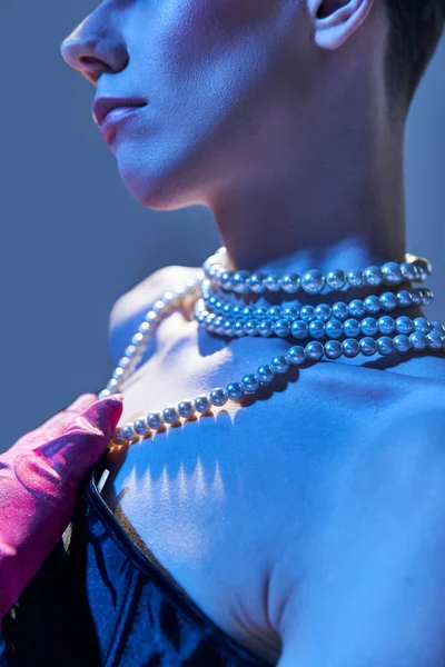 Primer plano y detalles, modelo genderfluid con collar de perlas posando en guante rosa sobre fondo azul - foto de stock