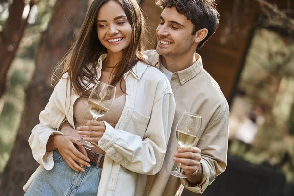 Homem positivo abraçando namorada com vinho perto de casa de férias borrada no fundo — Fotografia de Stock