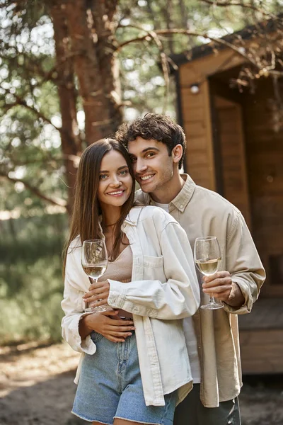 Lächelndes Paar umarmt sich mit Wein und blickt gemeinsam in die Kamera neben verschwommenem Ferienhaus — Stockfoto