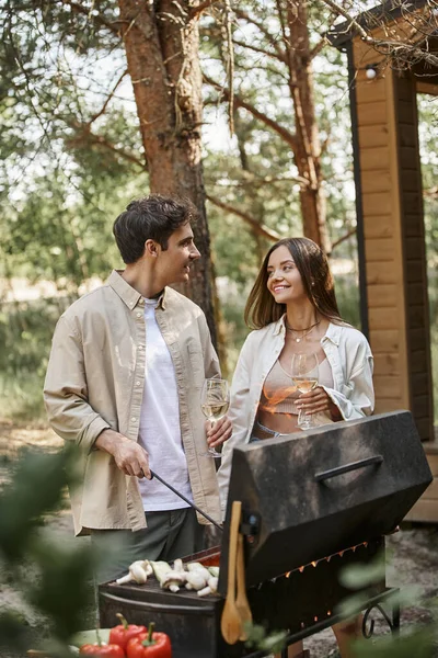 Morena homem cozinhar na grelha e falando com a namorada sorridente com vinho perto de casa de férias — Fotografia de Stock