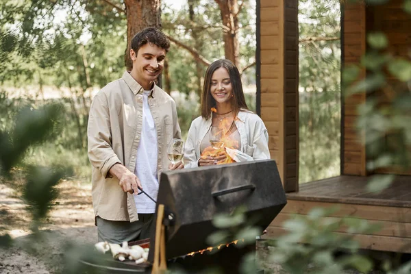 Smiling couple with wine cooking on grill while resting near vacation house at background — Stock Photo