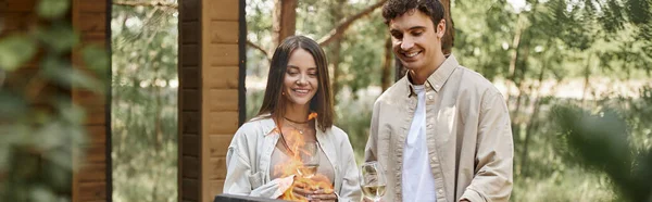 Mujer sonriente con vino cerca de novio y fuego en la parrilla durante el picnic cerca de la casa de vacaciones, pancarta - foto de stock