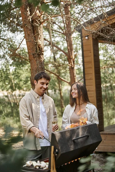 Coppia allegra con vino che cucina sulla griglia durante il picnic vicino alla casa estiva sullo sfondo — Foto stock