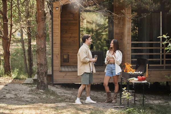 Casal positivo e elegante com vinho falando perto de churrasqueira e casa de férias em segundo plano — Fotografia de Stock