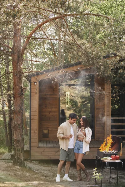Couple souriant et élégant avec vin étreignant près du barbecue pendant le pique-nique près de la maison de vacances en plein air — Photo de stock