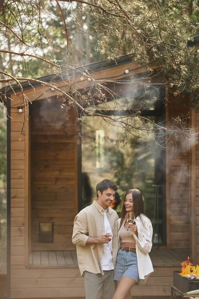 Cheerful and stylish couple holding wine and standing near grill with smoke and vacation house — Stock Photo