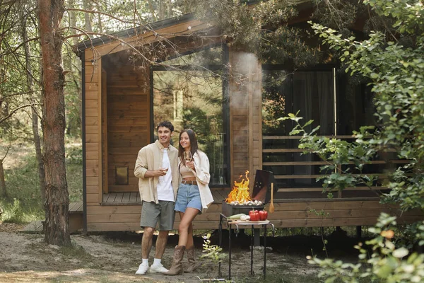 Positiva pareja romántica sosteniendo el vino y de pie cerca de la parrilla y la casa de vacaciones en el fondo - foto de stock