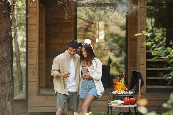 Alegre y elegante pareja sosteniendo el vino cerca de la parrilla y el humo con casa de vacaciones en el fondo - foto de stock