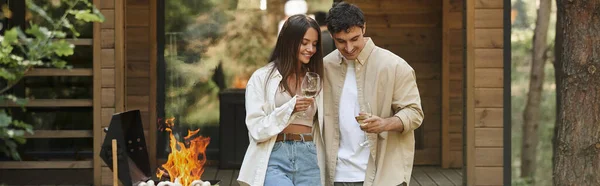 Positive couple holding wine near grill and blurred vacation house at background, banner — Stock Photo