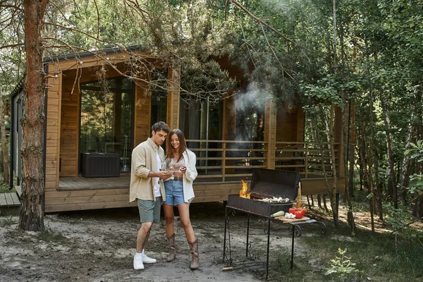 Smiling casal romântico brindar com vinho e pé perto de churrasqueira e casa de férias ao ar livre — Fotografia de Stock