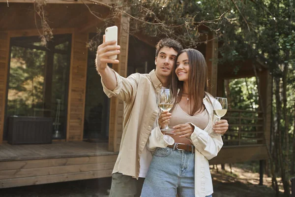 Elegante hombre tomando selfie en el teléfono inteligente con novia sosteniendo el vino cerca de la casa de verano en el fondo - foto de stock