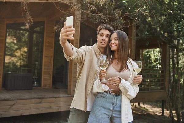Stylish man taking selfie on smartphone with girlfriend holding wine near summer house at background — Stock Photo