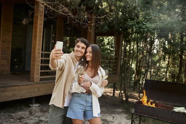 Casal sorridente com vinho tirando selfie no smartphone perto de churrasco e casa de férias no fundo — Fotografia de Stock