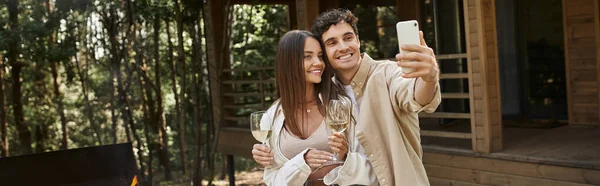 Coppia sorridente con selfie di vino su smartphone vicino bbq e casa estiva sullo sfondo, banner — Foto stock