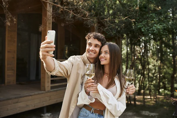 Lächelnde junge Frau hält Wein in der Hand, während ihr Freund in der Nähe des Ferienhauses ein Selfie mit dem Smartphone macht — Stockfoto