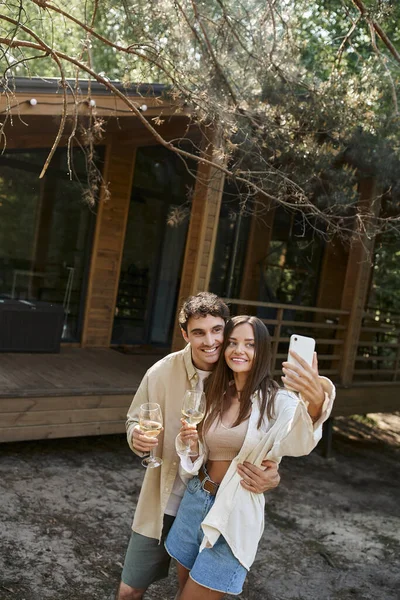 Mujer sonriente sosteniendo vino y tomando selfie en el teléfono inteligente con el novio cerca de la casa de vacaciones - foto de stock