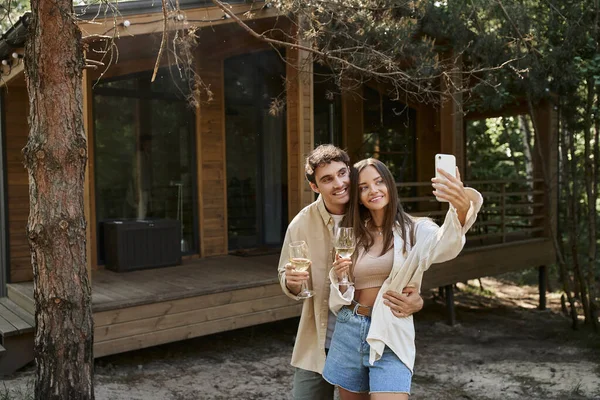 Sorrindo homem abraçando namorada e tomando selfie com namorada segurando vinho perto de casa de férias — Fotografia de Stock