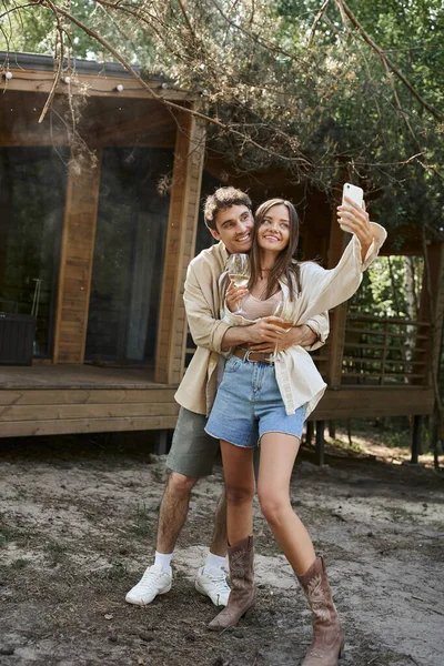 Homme souriant étreignant petite amie avec du vin prendre selfie sur smartphone près de la maison de vacances en plein air — Photo de stock