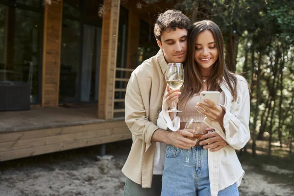 Stylish man hugging girlfriend with wine using smartphone near vacation house at background — Stock Photo
