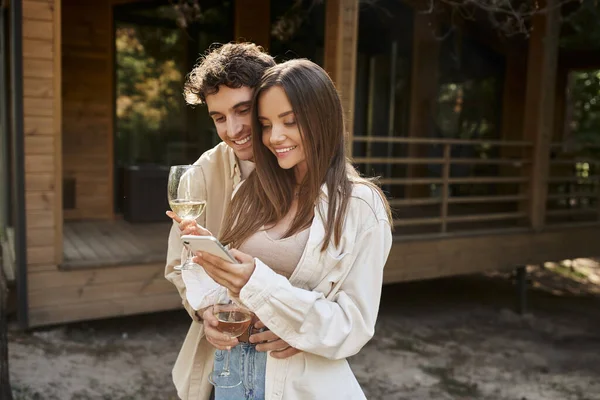 Улыбающаяся романтическая пара с вином с помощью смартфона возле размытого летнего домика на заднем плане — стоковое фото