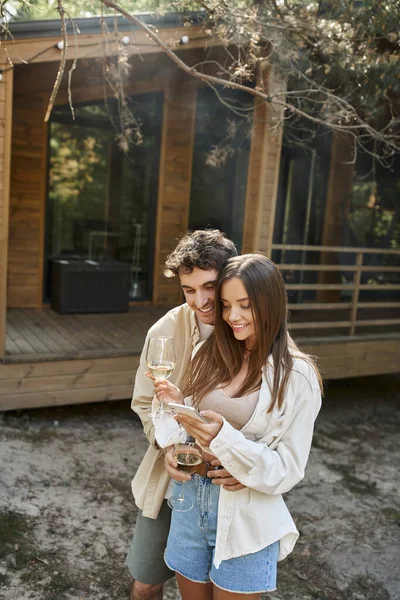 Sonriente y elegante pareja con vino utilizando el teléfono móvil cerca de la casa de vacaciones borrosa en el fondo — Stock Photo
