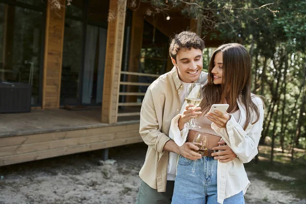 Sonriente hombre con vino abrazando novia utilizando el teléfono móvil cerca de la casa de vacaciones en segundo plano — Stock Photo