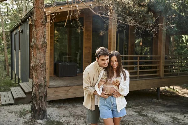 Romantisches Paar mit Wein lächelt während Smartphone in der Nähe von Ferienhaus im Freien — Stockfoto