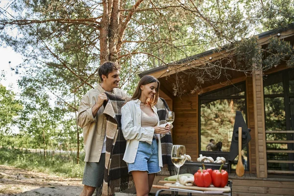 Lächelnder Mann hält Decke neben Freundin mit Wein, Grill und Ferienhaus im Hintergrund — Stockfoto