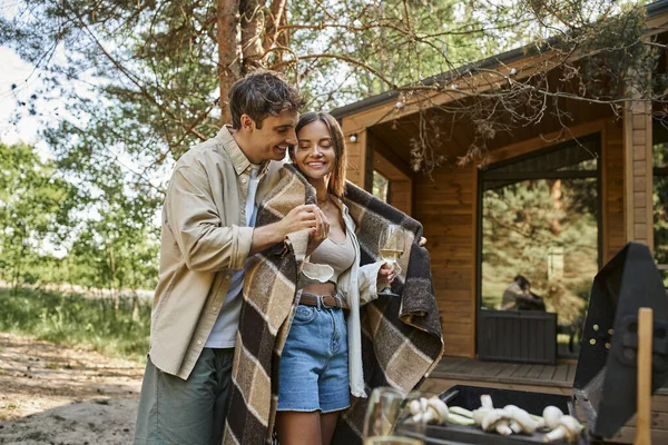 Cheerful man holding blanket near girlfriend with wine, bbq and summer house at background — Stock Photo