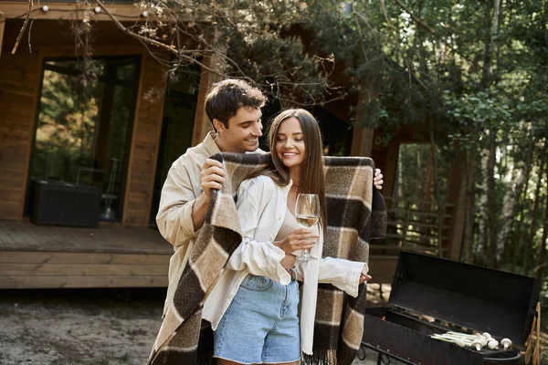 Homme tenant couverture près de petite amie avec du vin, grill et maison de vacances à l'arrière-plan — Photo de stock