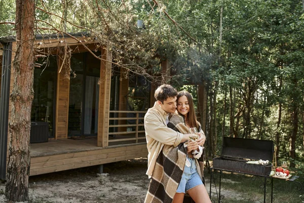 Homme embrassant petite amie souriante avec couverture et vin tout en se tenant près du gril et de la maison de vacances — Photo de stock