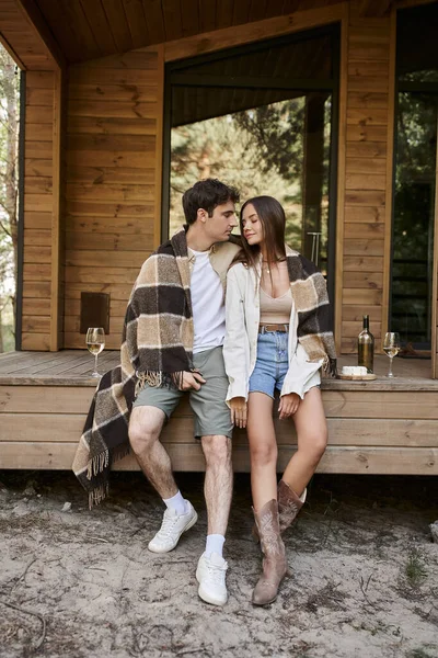 Romantisches Paar in Decke bei Wein und Käse sitzend mit Ferienhaus im Hintergrund — Stockfoto
