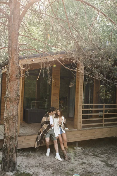 Улыбающаяся пара в одеяле держит вино возле сыра и летний дом под деревьями на открытом воздухе — стоковое фото