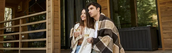 Pareja sonriente en manta sosteniendo el vino y mirando lejos cerca de la casa de vacaciones al aire libre, pancarta - foto de stock