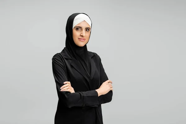 Портрет уверенной мусульманки-предпринимательницы в хиджабе и черной куртке, позирующей со сложенными руками на сером — стоковое фото