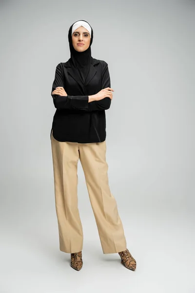 Стильная мусульманская бизнесвумен в пиджаке, брюках и хиджабе позирует со сложенными руками на сером, полной длины — стоковое фото