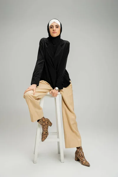 Businesswoman in muslim hijab and trendy casual attire sitting on high stool on grey, full length — Stock Photo