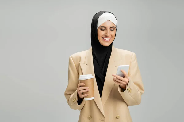 Mulher de negócios muçulmana motivada em hijab e blazer, com smartphone e café para ir em cinza — Fotografia de Stock