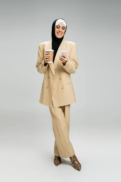 Exitosa mujer de negocios musulmana en traje hijab y beige sosteniendo taza de papel y teléfono móvil en gris - foto de stock