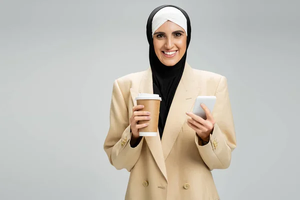 Feliz mujer de negocios musulmana en blazer beige y hijab sosteniendo café y teléfono inteligente en gris - foto de stock