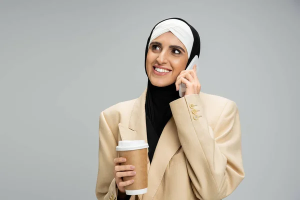 Alegre y elegante mujer de negocios musulmana sosteniendo café para ir y hablar en el teléfono inteligente en gris - foto de stock