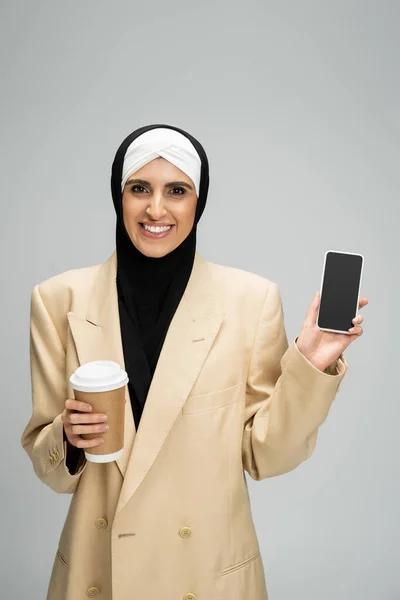 Улыбающаяся, модная мусульманка-бизнесмен с кофе идет показывать смартфон с чистым экраном на сером — стоковое фото