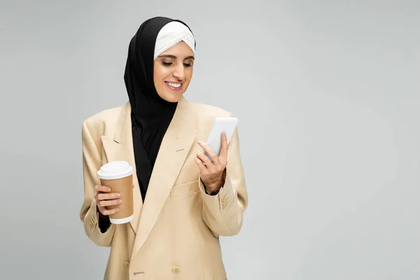 Mujer de negocios musulmana feliz y de moda con red de bebidas para llevar en el teléfono inteligente en gris - foto de stock
