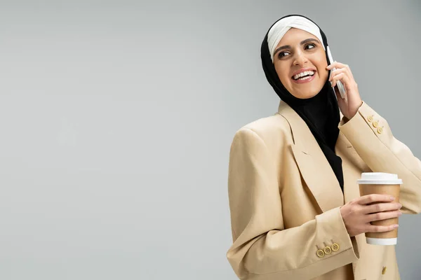 Elegante mujer de negocios musulmana con bebida para llevar riendo y hablando en el teléfono móvil en gris, - foto de stock
