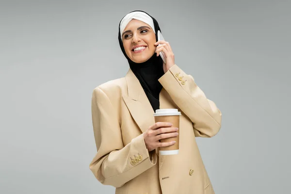 Alegre, mujer de negocios musulmana de moda con café para ir a hablar en el teléfono inteligente, mirando hacia otro lado en gris - foto de stock