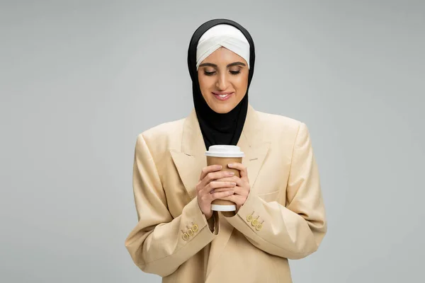 Pleased middle eastern woman in muslim hijab and beige blazer holding coffee to go on grey — Stock Photo