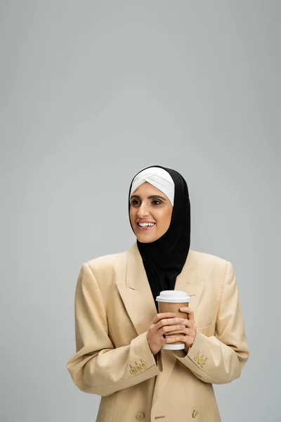 Mujer musulmana positiva en chaqueta elegante y hijab sosteniendo taza de papel y mirando hacia otro lado en gris - foto de stock