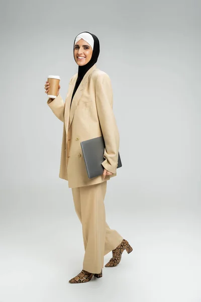 Mulher muçulmana bem sucedida empresária com copo de papel, laptop e sorriso radiante em cinza, comprimento total — Fotografia de Stock
